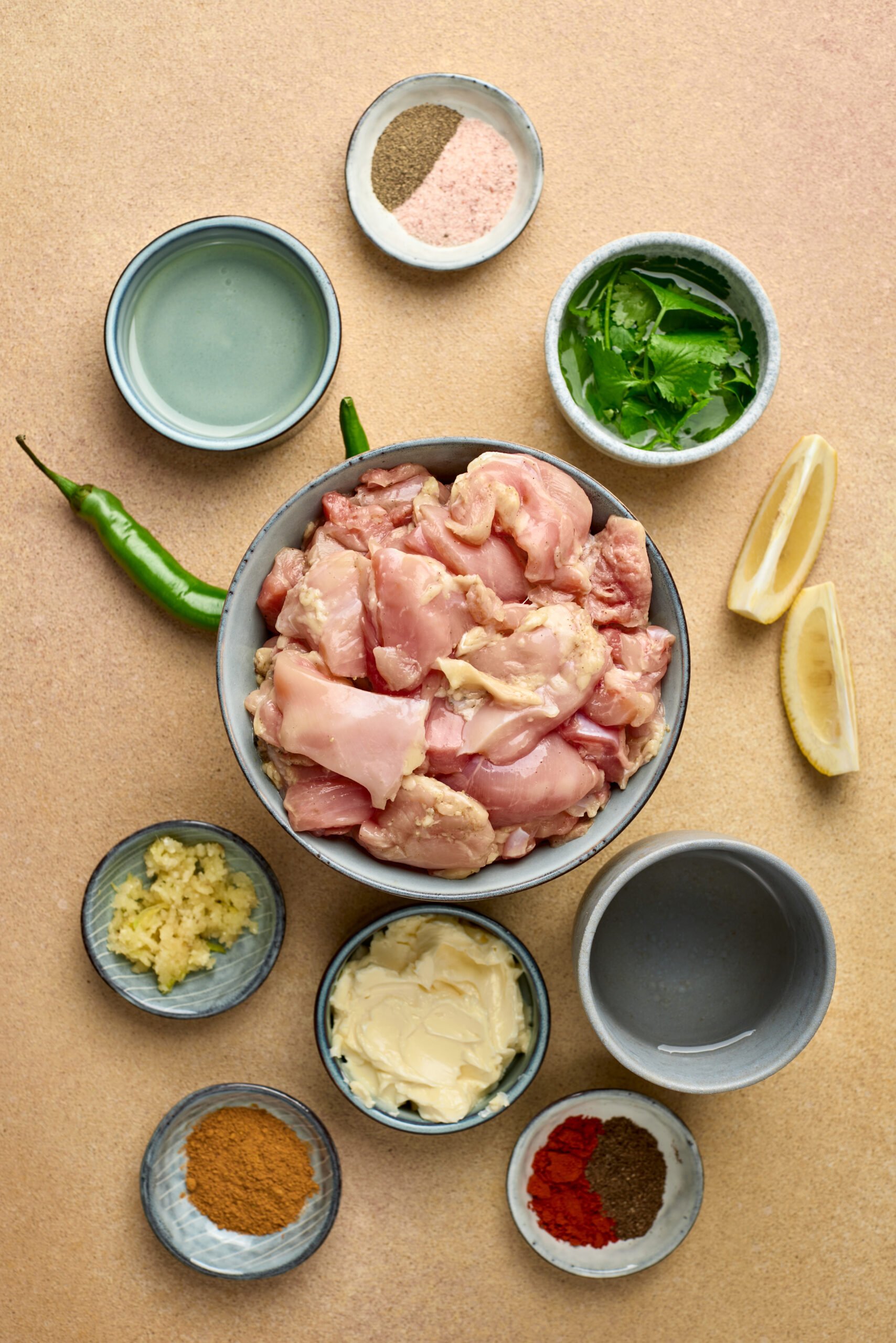 Ingredients for garlic butter chicken including raw chicken pieces, garlic cloves, butter, cumin, cream, and fresh parsley arranged on a kitchen counter