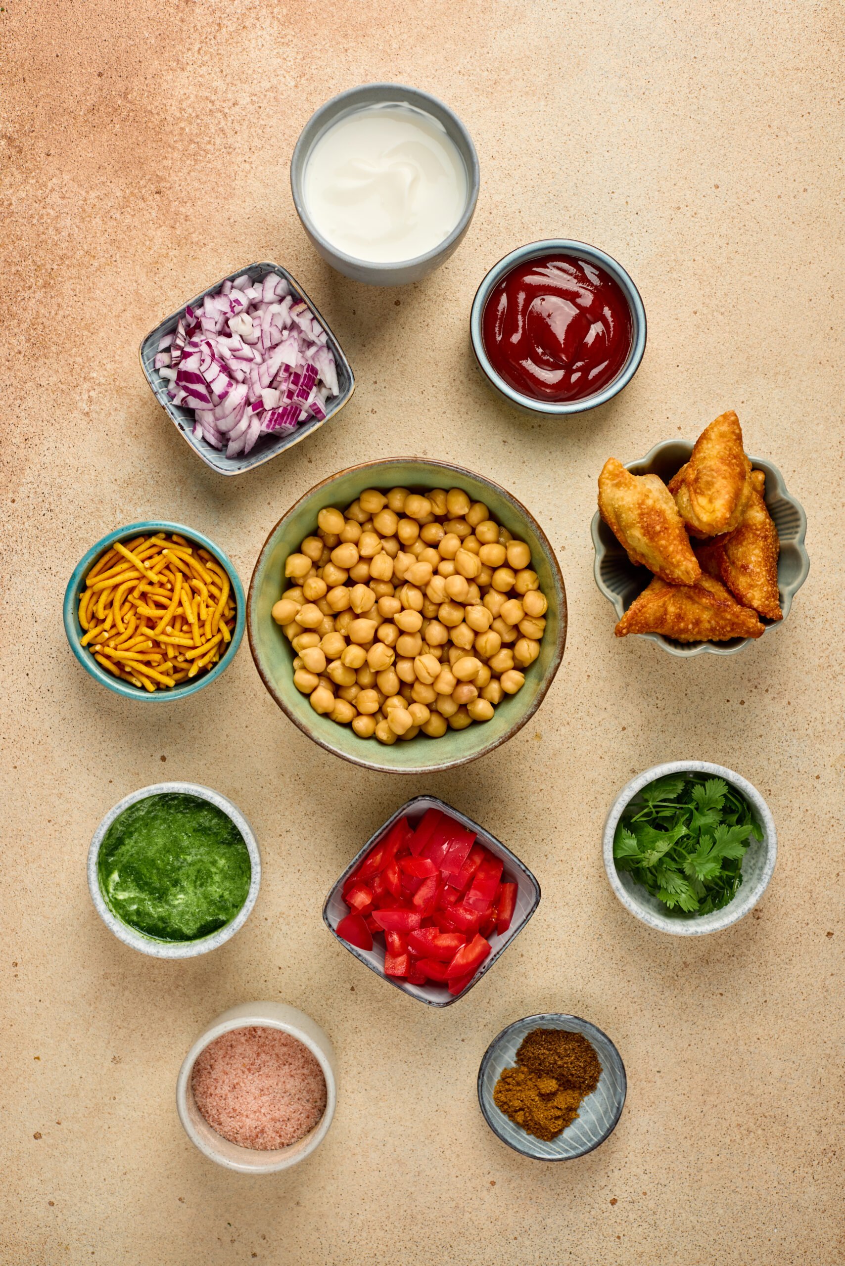 Ingredients for Samosa Chaat including samosas, yogurt, chickpeas, tamarind chutney, green chutney, onions, tomatoes, and sev arranged on a counter.