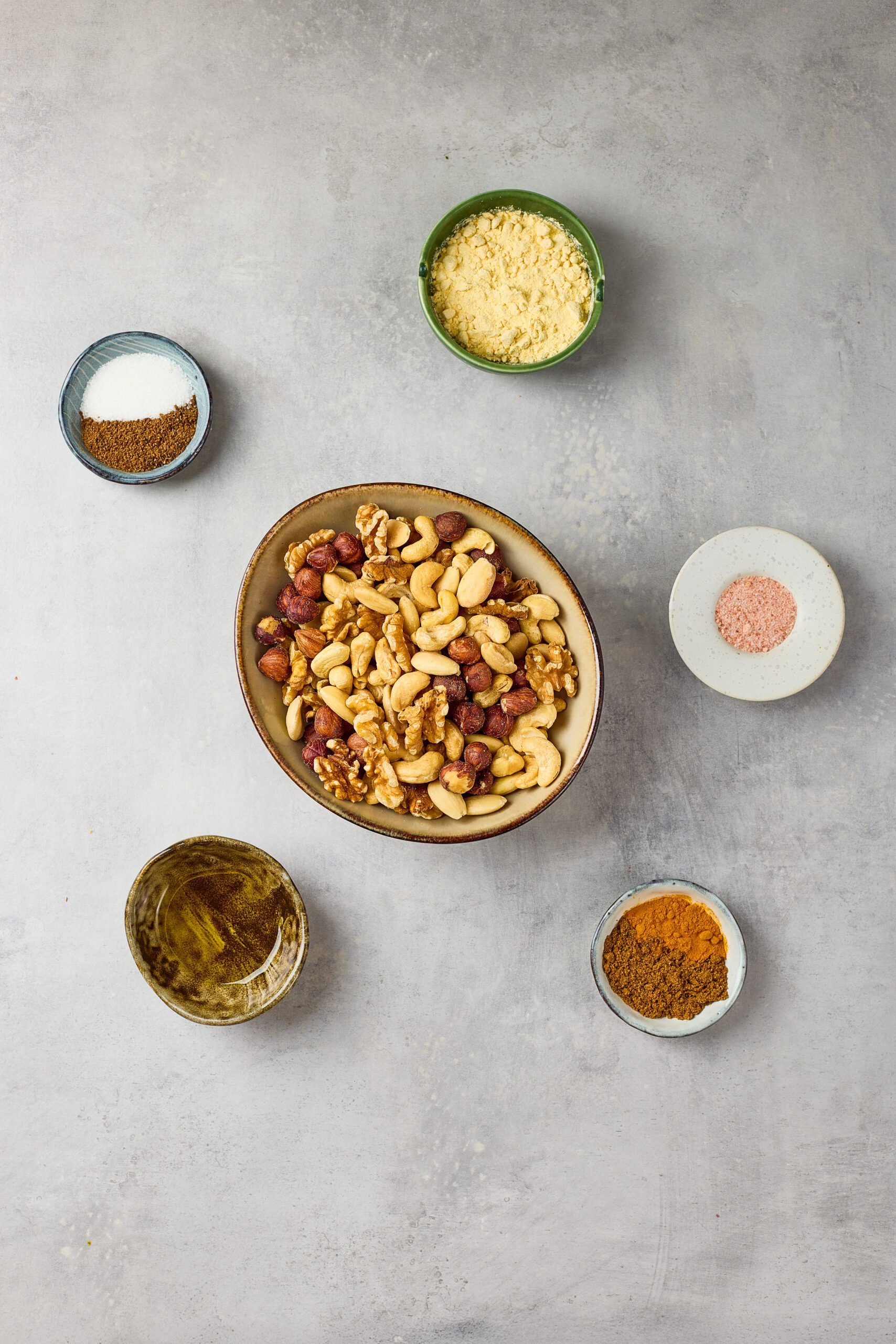 Ingredients for Spiced Nuts including mixed nuts, olive oil, honey, and spices like chili powder, cumin, and cinnamon arranged on a counter.