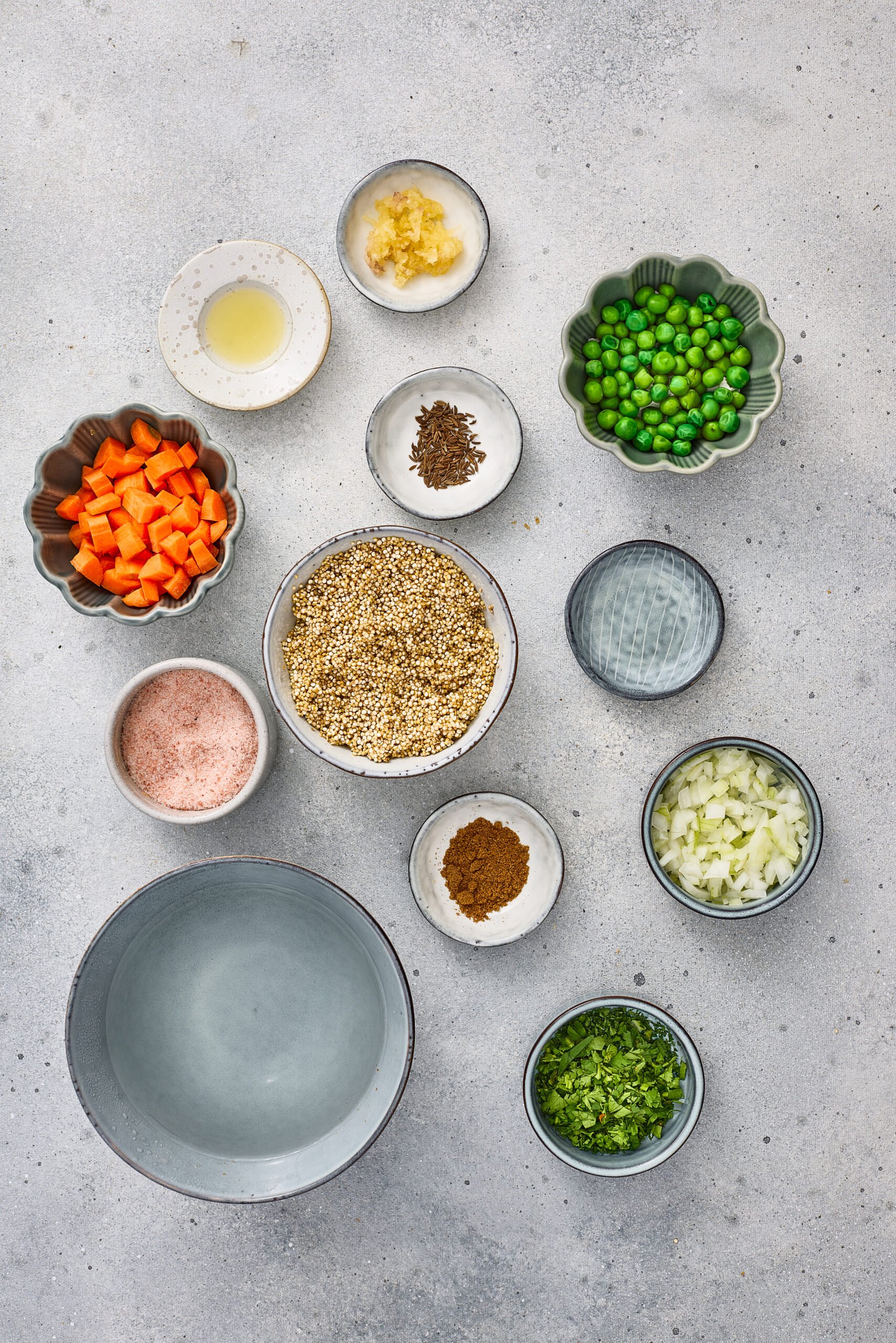 Ingredients for Quinoa Pulao displayed in bowls: quinoa, carrots, peas, onions, cumin seeds, garam masala, ginger, coriander, salt, and water.